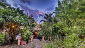 Un jardín tropical con una estructura de techo de paja, un área de comedor y un sendero que conduce a ella.