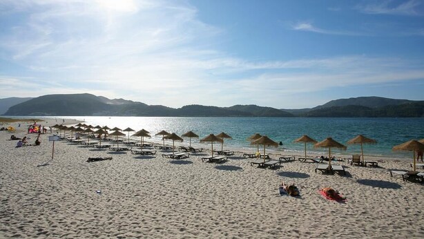 Une plage bordée de rangées de chaises longues et de parasols, un ciel dégagé et des montagnes en arrière-plan.