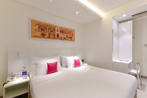 Habitación de hotel con una cama grande, paredes blancas, un cuadro sobre la cama, una mesita de noche con un teléfono y una botella, y un pequeño escritorio con una silla.
