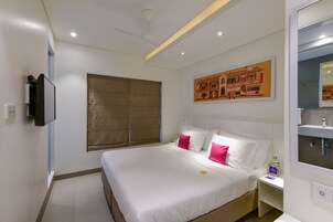 Habitación de hotel con cama, mesita de noche, televisor, ventilador de techo, ventana con persianas y un cuadro colgado en la pared.