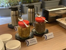 Una barra de desayuno con botes de copos de maíz y chocos, un tostador y tazas térmicas.