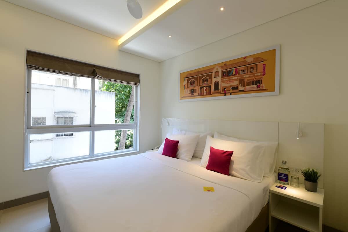 A hotel room with a large bed, a window with blinds, a wall-mounted painting, and a bedside table with a plant and a bottle.
