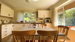 Een keuken met houten kastjes, een eettafel met stoelen, een magnetron en een raam met gordijnen.