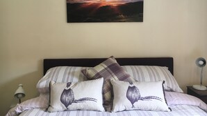 Een slaapkamer met een bed, kussens, een nachtkastje, een lamp en een schilderij van een zonsondergang aan de muur.