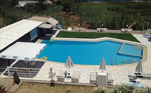 A swimming pool with lounge chairs and umbrellas, surrounded by a grassy area and buildings. - Andysgardens (Platanias)