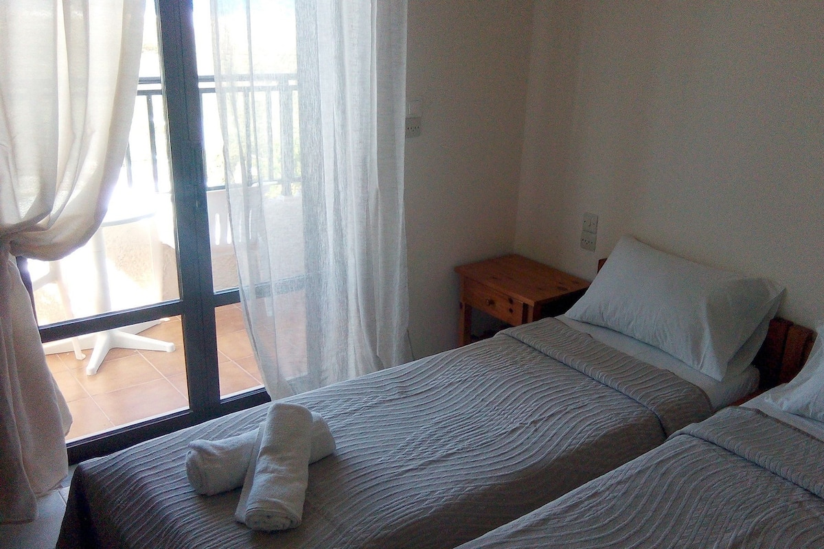 A bedroom with two beds, a wooden nightstand, and a window with sheer curtains.