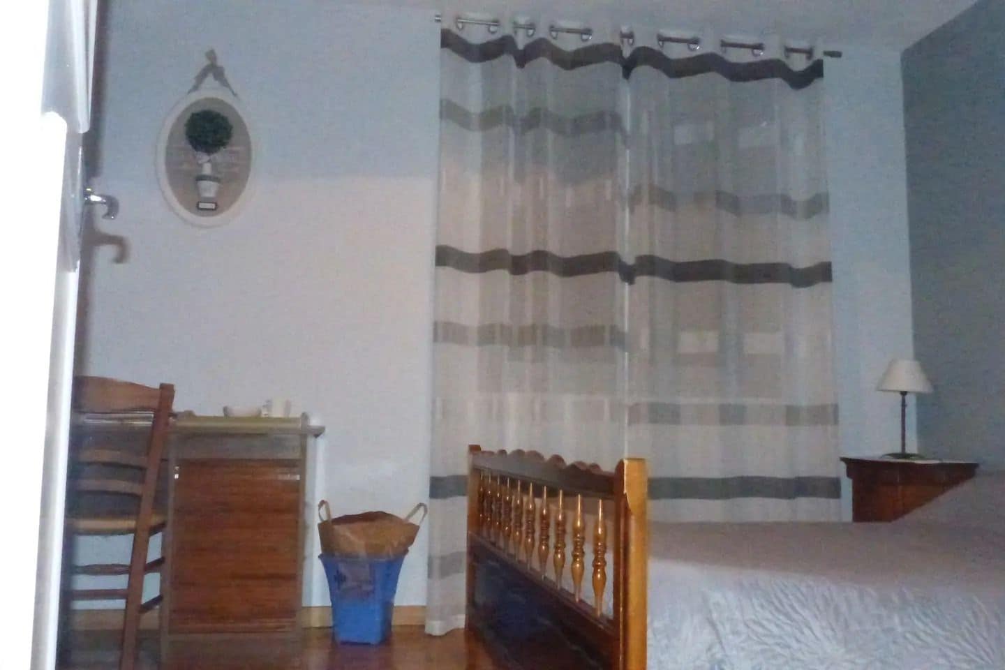 A bedroom with a bed, a wooden chair, a desk, a blue trash bin, a lamp, and a window with sheer curtains.