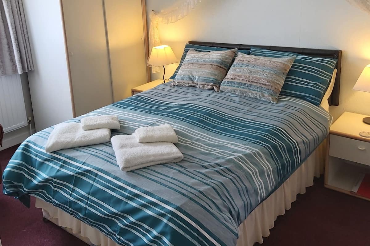 En snyggt bäddad säng med randigt sänglinne och två handdukar, ett sängbord med en lampa och en garderob i ett hotellrum.