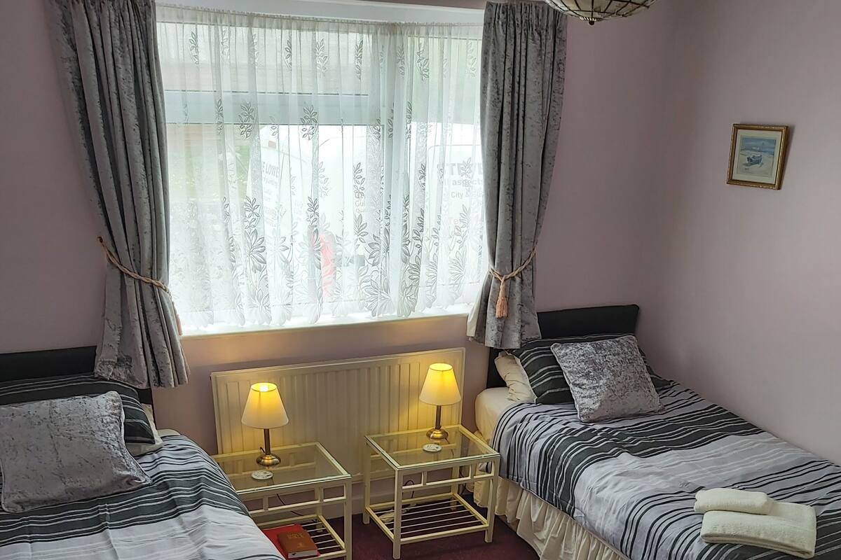 Ett hotellrum med två separata sängar, ett fönster med tunna gardiner, ett nattduksbord med en lampa och ett litet element.