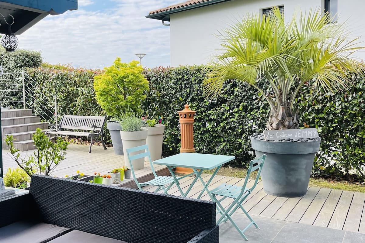 A patio area with a wicker sofa, a table, and chairs, surrounded by potted plants and a white building in the background.