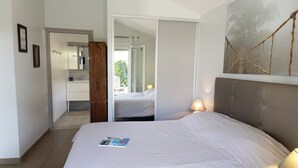 Quarto com cama, mesa de cabeceira, espelho, porta para outro cômodo e uma obra de arte na parede.
