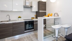 A modern kitchen with dark wood cabinets, a stainless steel range hood, and a white countertop. There are two bar stools and a small white island with a sink. - Apartments Nada (Dubrovnik)