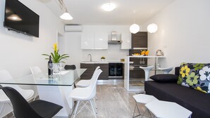 A modern kitchen with white cabinets, a built-in oven, and a dining area with a glass table and white chairs. - Apartments Nada (Dubrovnik)