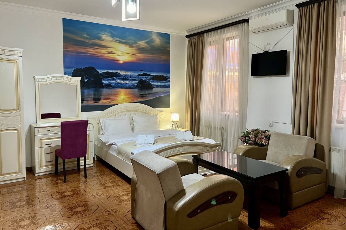 A hotel room with a bed, a desk, a chair, a TV, and a painting of a sunset over the ocean.