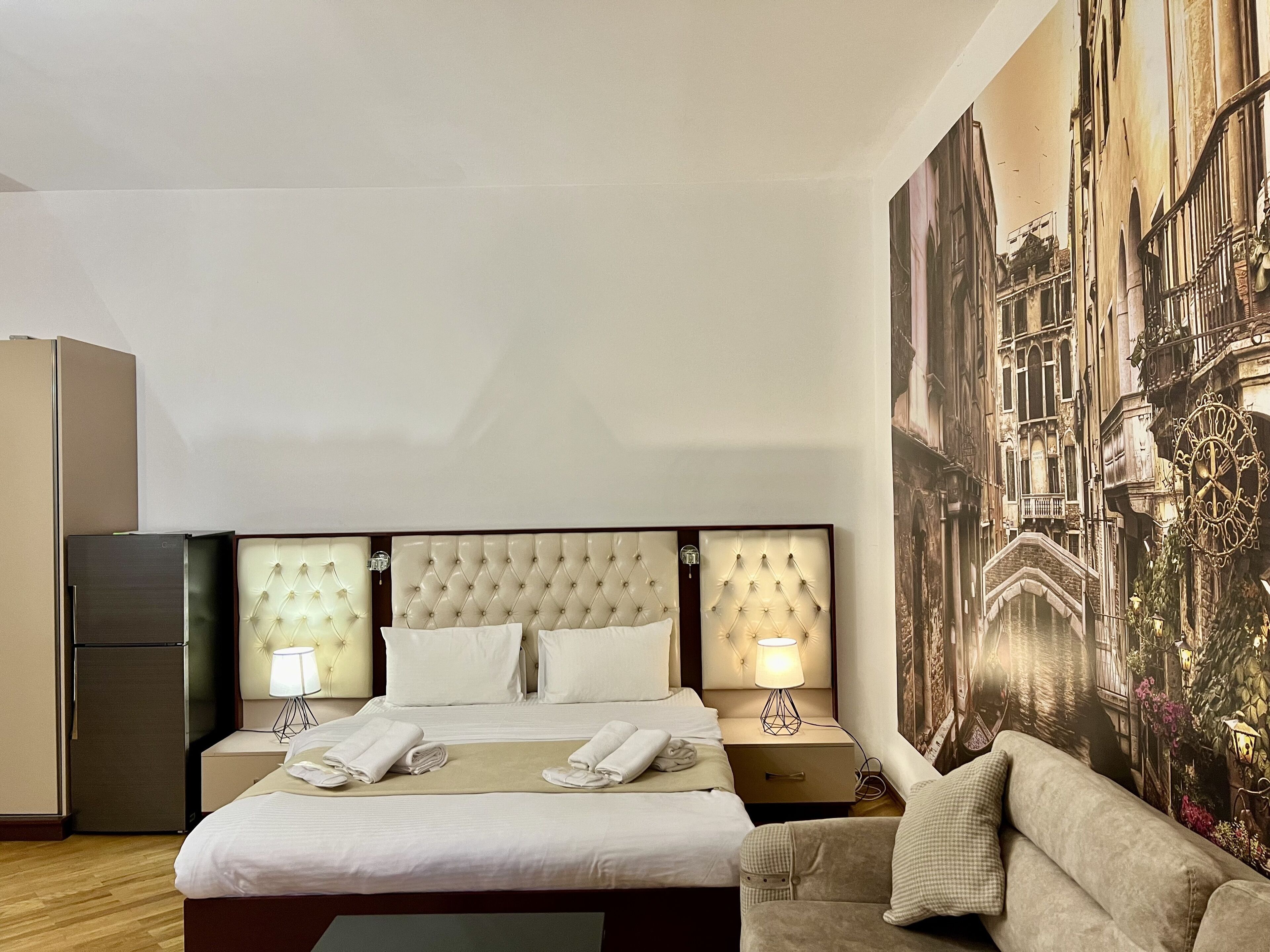 A hotel room with a large bed, two bedside tables with lamps, a sofa, and a wall mural of a cityscape.