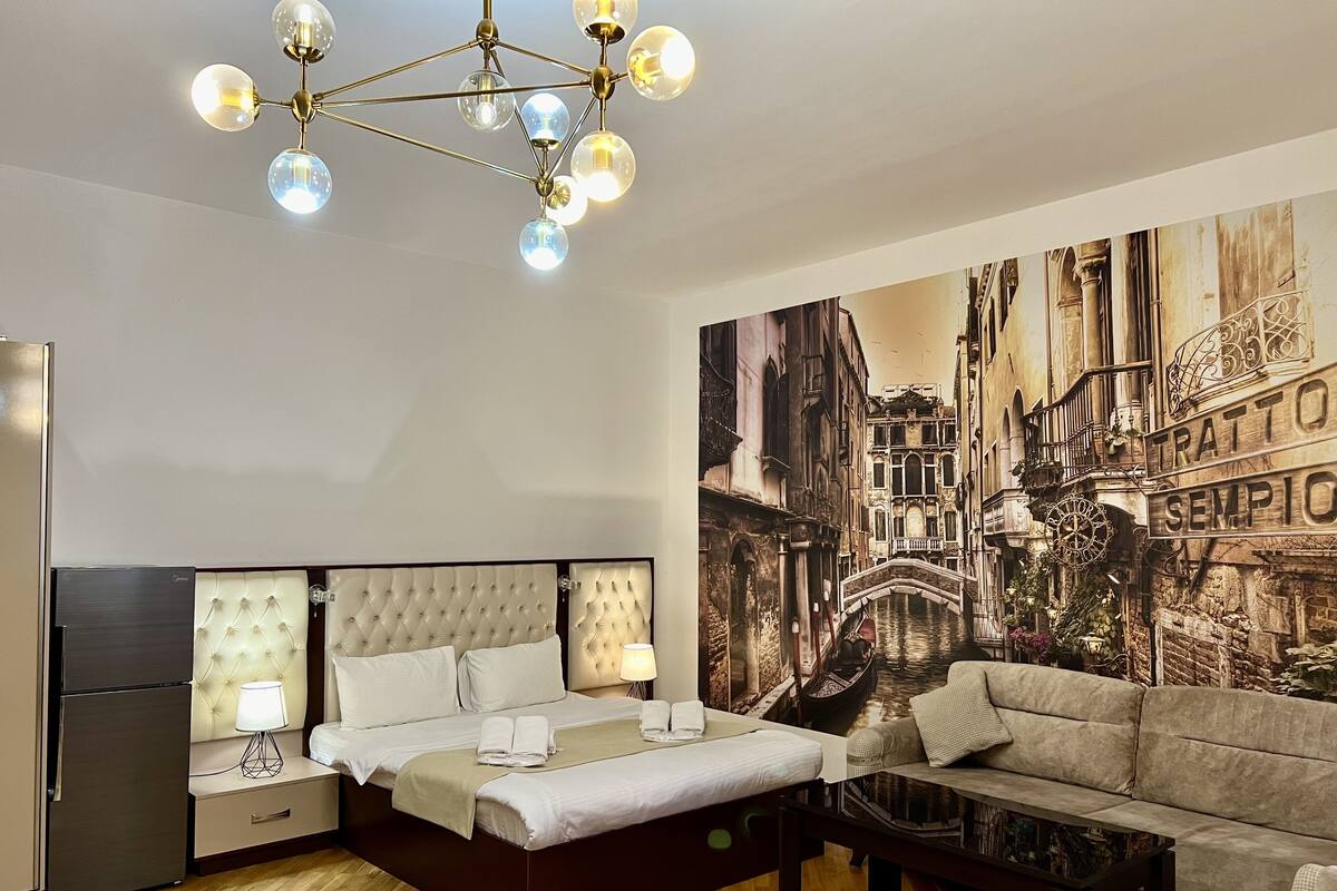 A hotel room with a bed, a sofa, a glass coffee table, and a mural of a cityscape.