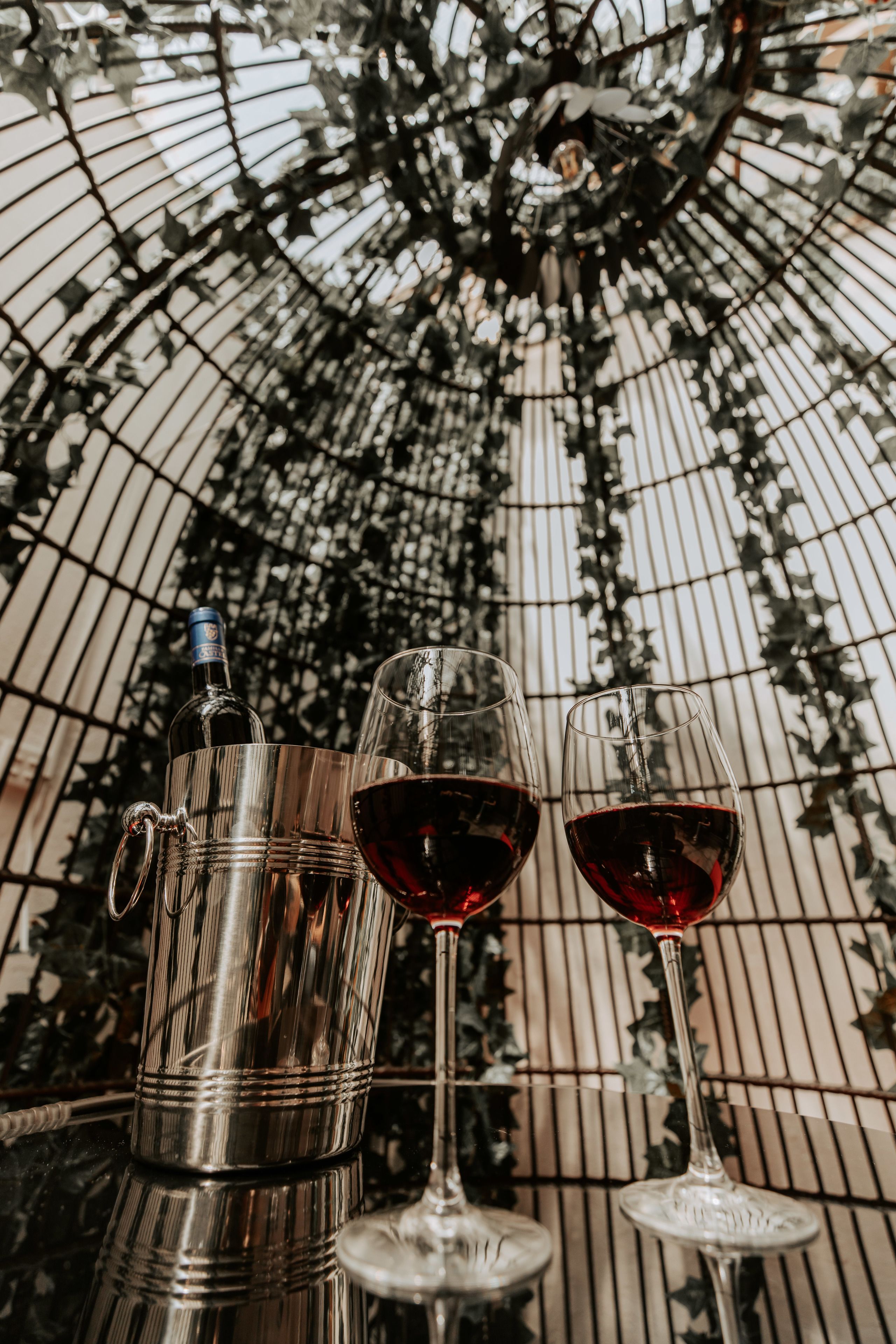 A metallic wine cooler with a bottle and two wine glasses on a table under a decorative umbrella.