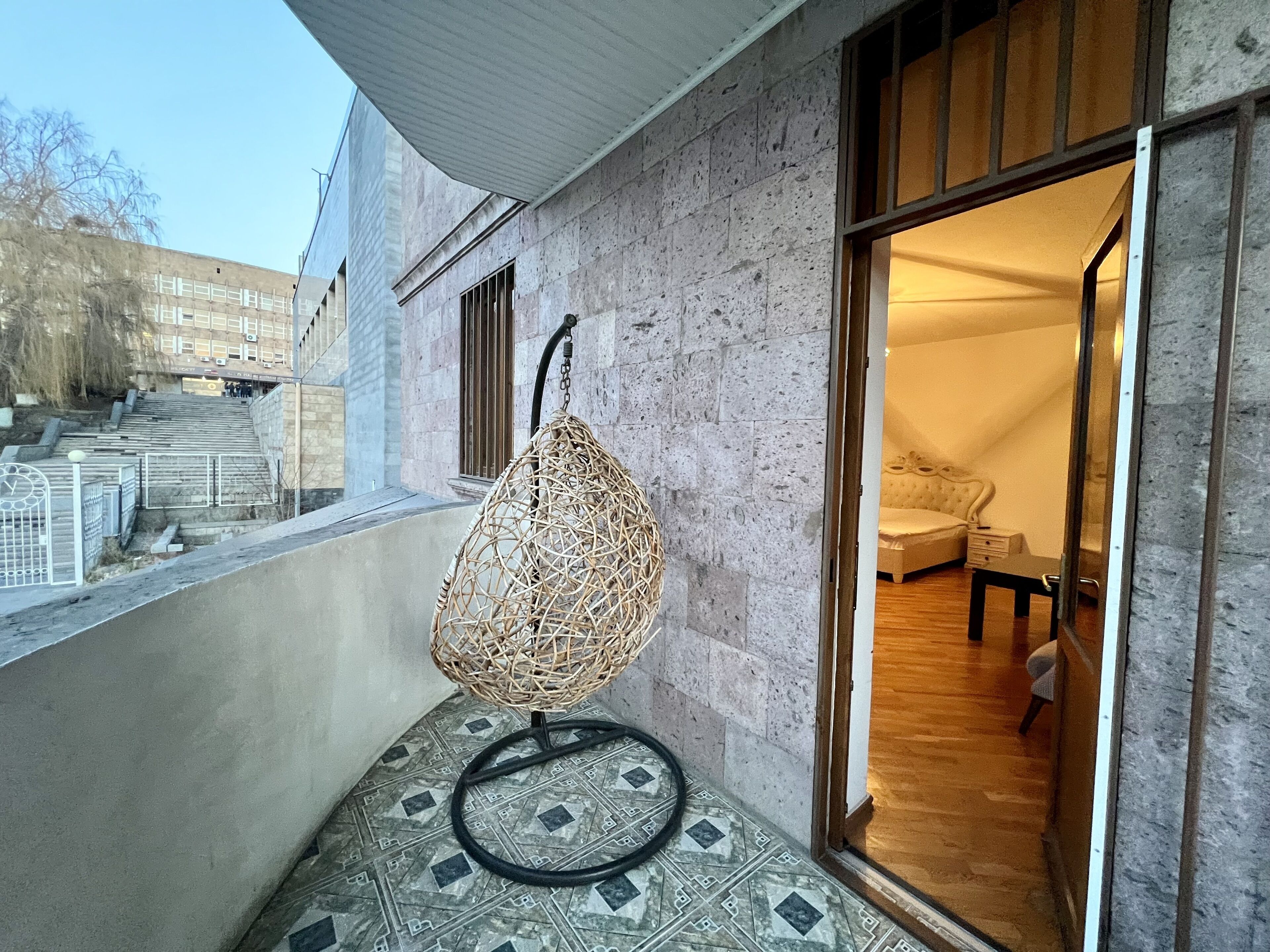 A wicker chair on a balcony with patterned tiles, a view of a building with barred windows, and a glimpse of a room with a bed and a table.