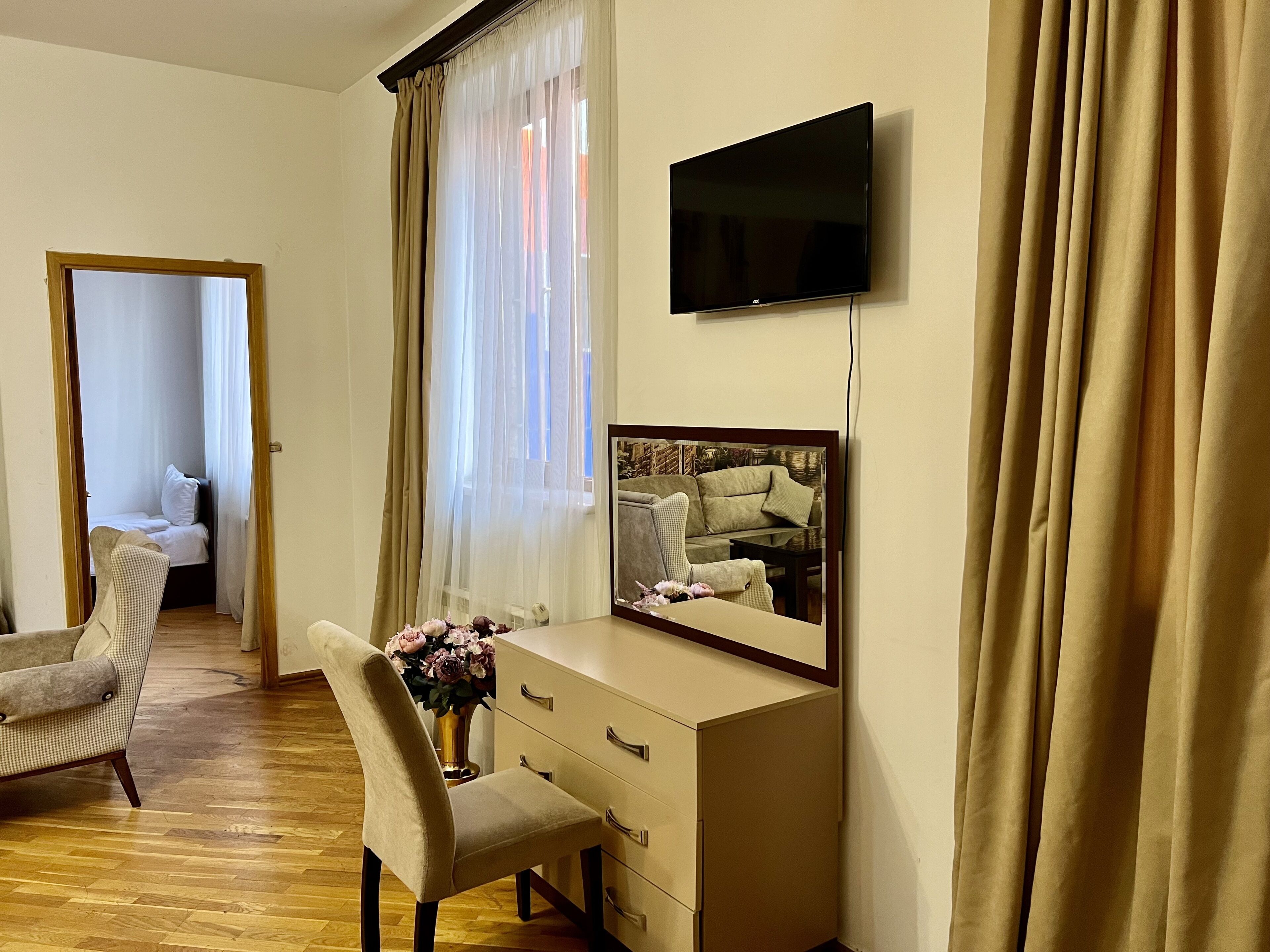 A hotel room with a bed, a desk with drawers, a chair, a television mounted on the wall, and a large window with curtains.