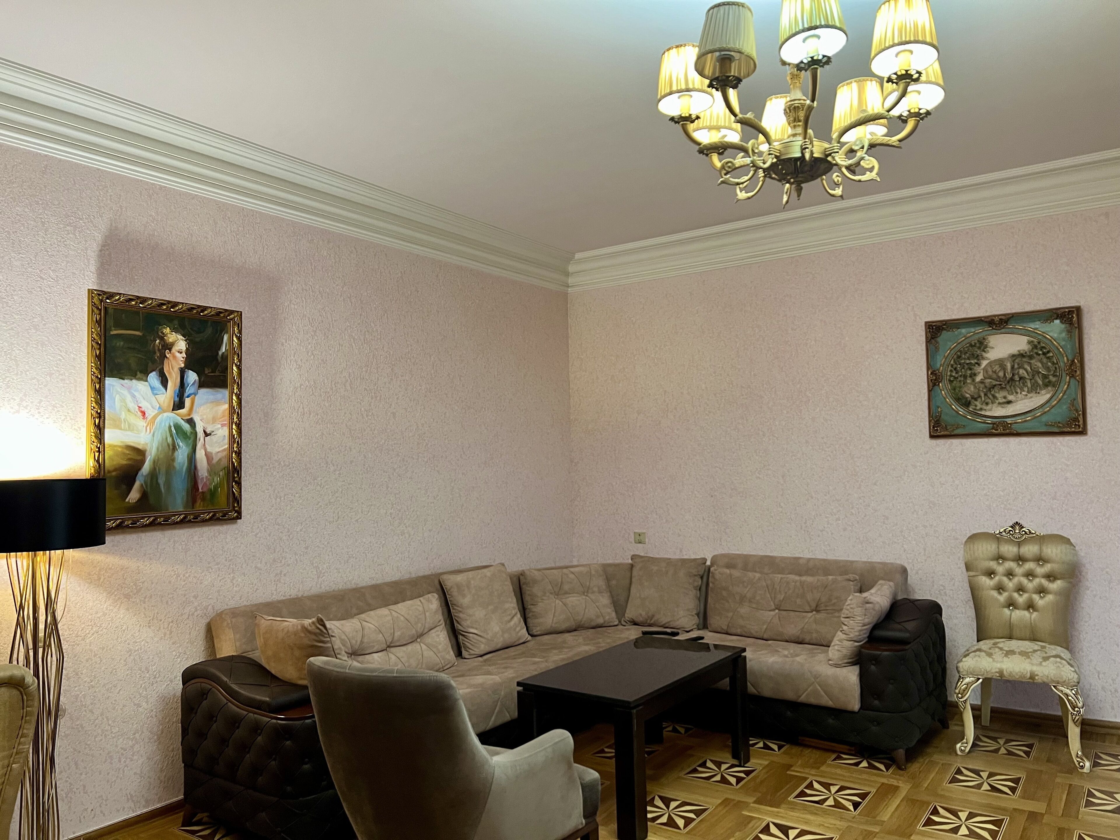 A corner room with a beige sectional sofa, a black armchair, a small table, a decorative chair, a chandelier, and two wall-mounted paintings.