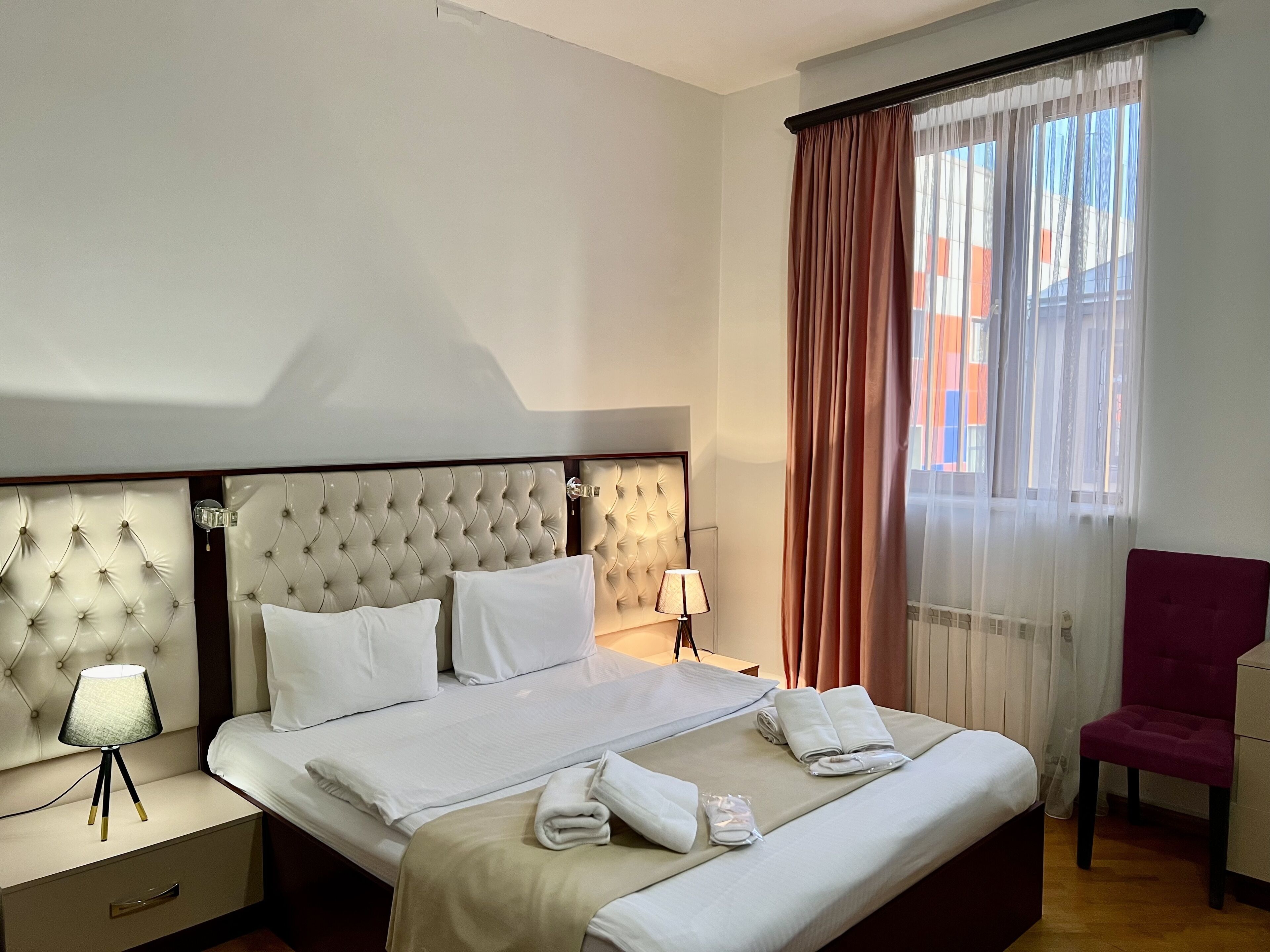 A hotel room with a large bed, a nightstand with a lamp, a chair, and a window with curtains.