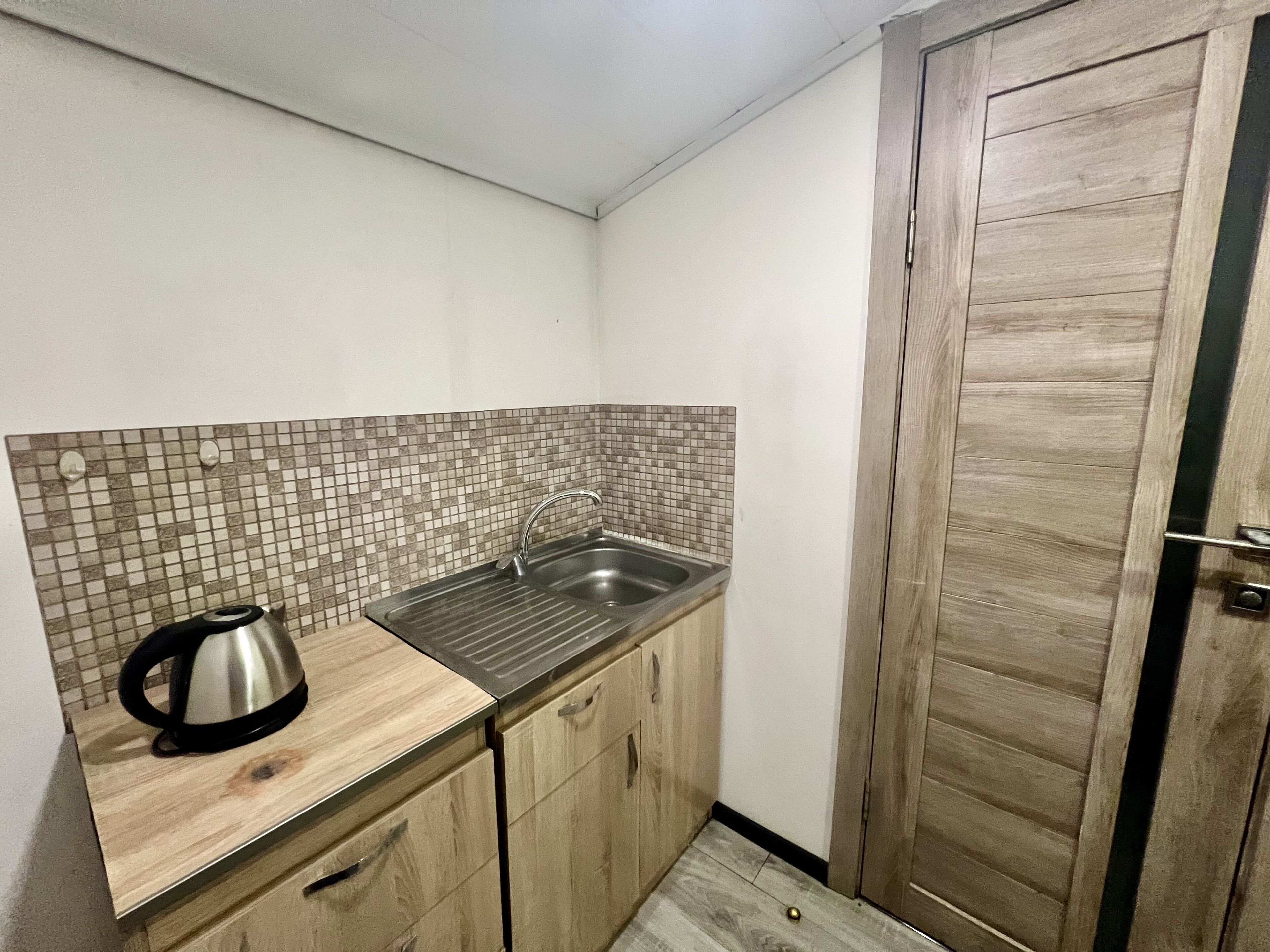A small kitchen with a tiled backsplash, a stainless steel sink, wooden cabinets, and a kettle on the counter.