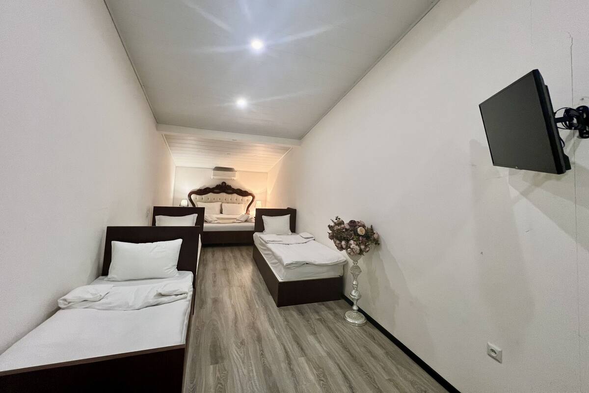 A narrow hotel room with two single beds, a flat-screen TV mounted on the wall, and a small bedside table with a vase of flowers.