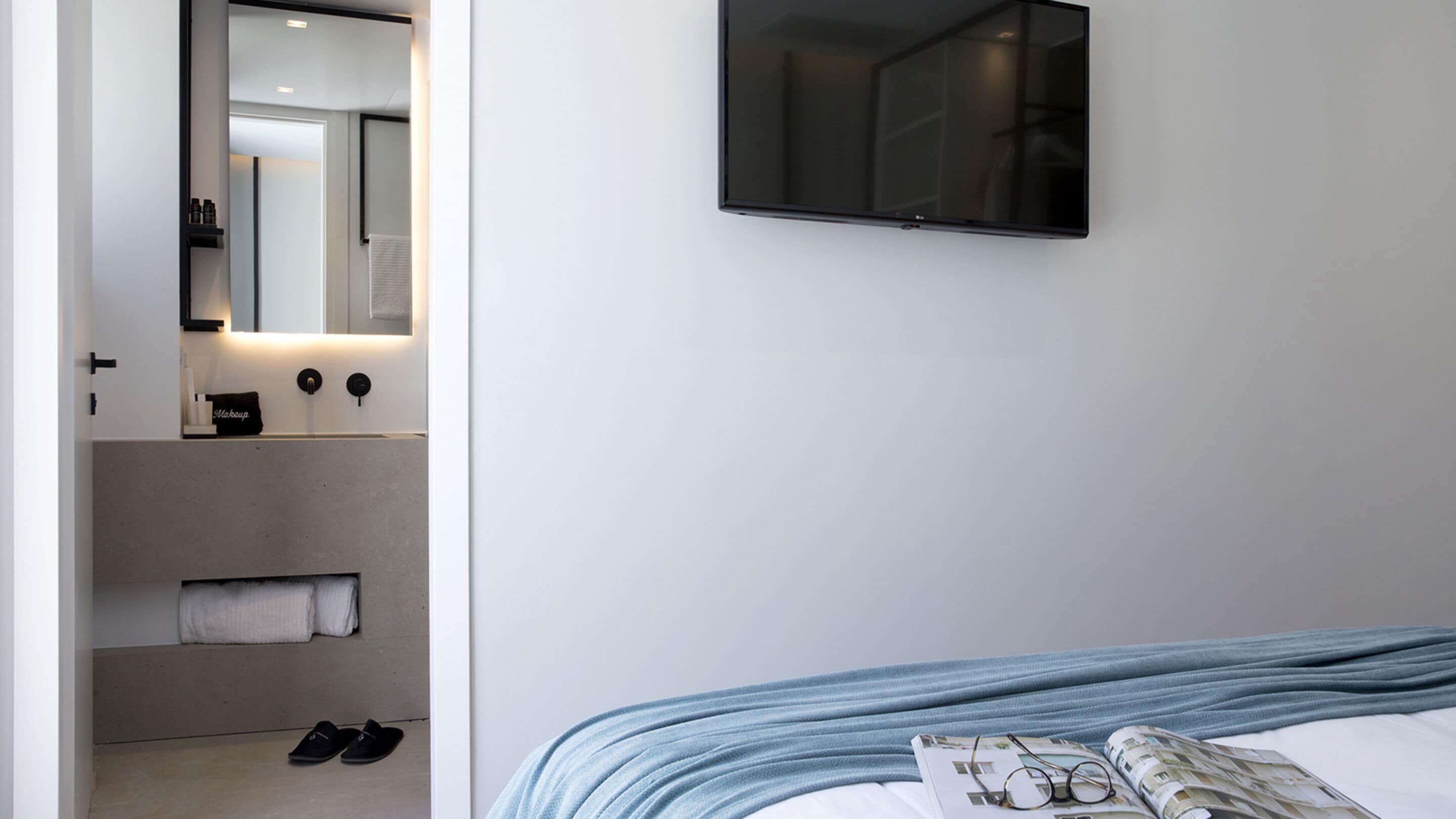 A modern hotel room with a bed, a mounted TV, a bathroom with a mirror, and a towel rack.