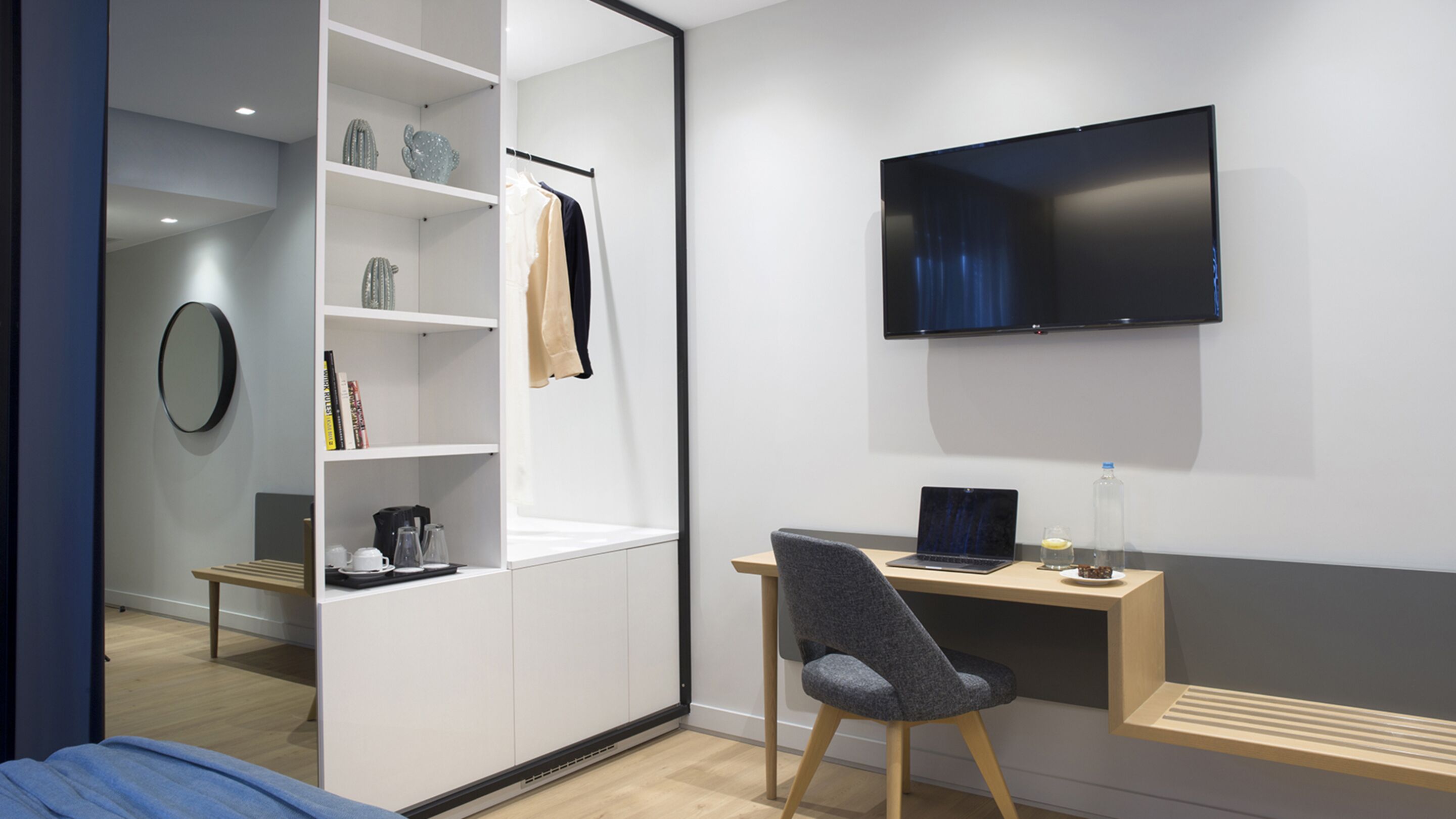 A modern bedroom with a bed, a desk with a laptop, a chair, a TV mounted on the wall, and a wardrobe with shelves.