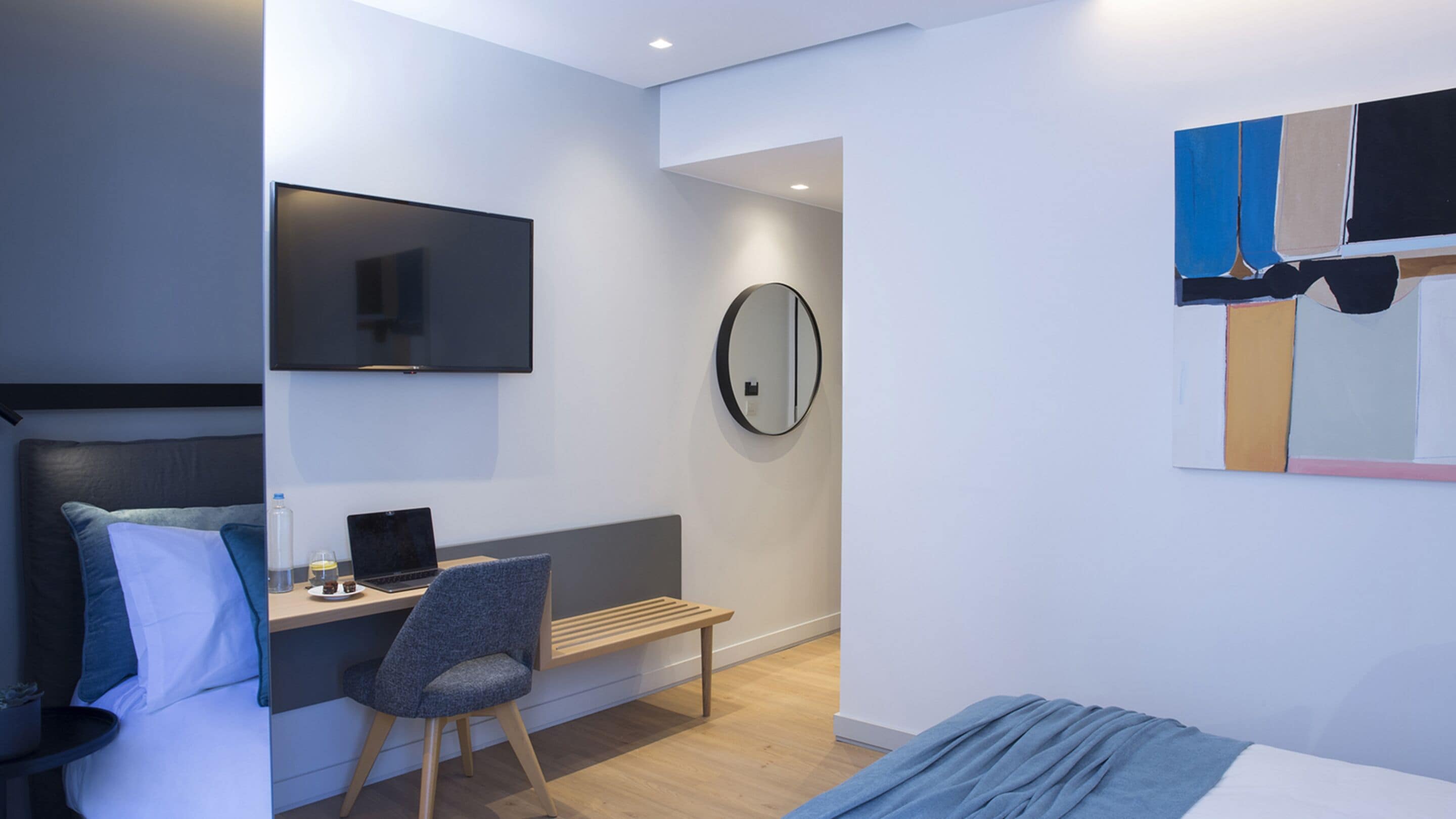 Una habitación de hotel moderna con una cama, un escritorio con una computadora portátil, una silla, un televisor y un espejo.
