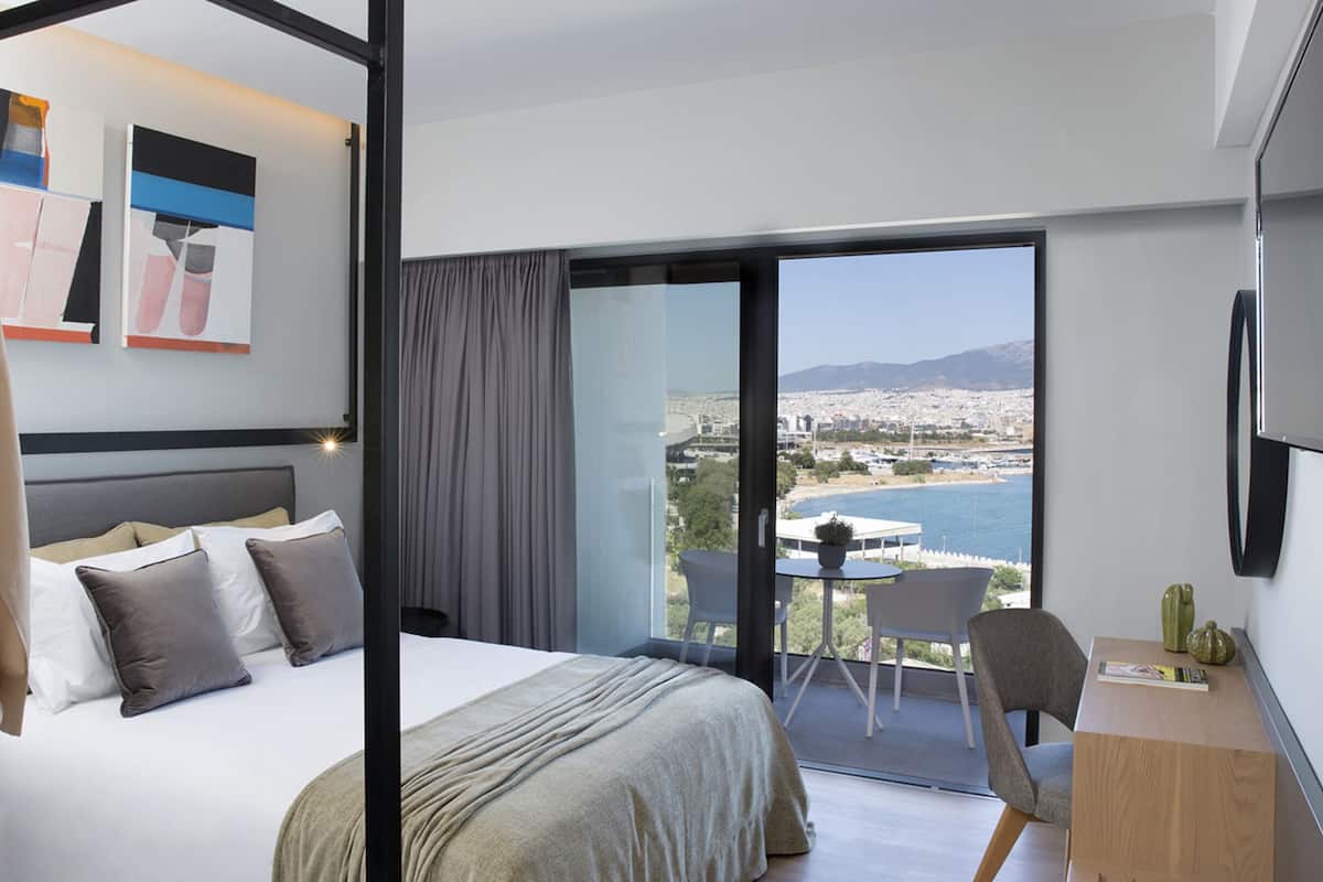 Una habitación de hotel moderna con una cama grande, un balcón con vistas al mar y a la ciudad, y un escritorio con una silla.