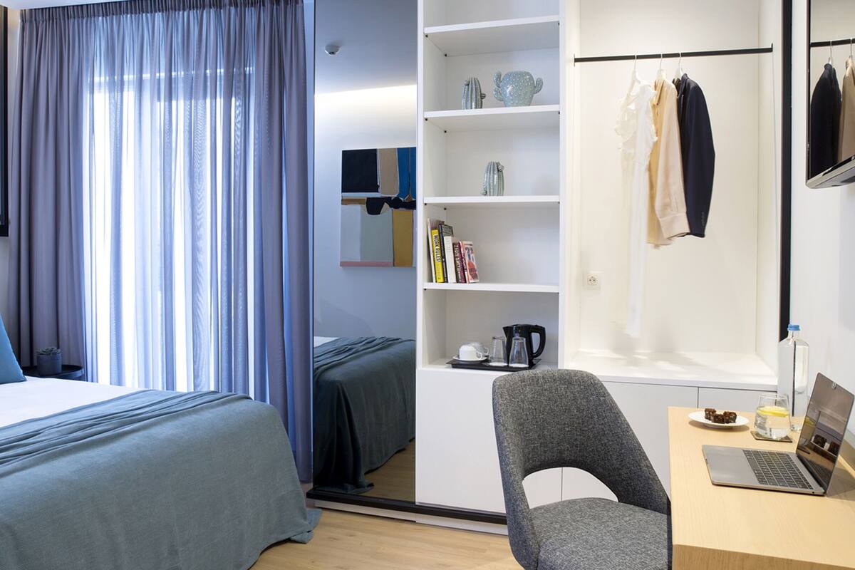 Una habitación de hotel con cama, escritorio con computadora portátil, silla, estantería y armario con ropa colgada.