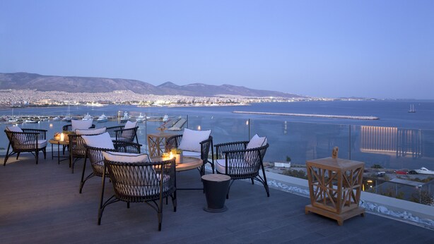 Un área de estar al aire libre con sillas de mimbre y una mesa, con vista a un puerto deportivo y al paisaje urbano.