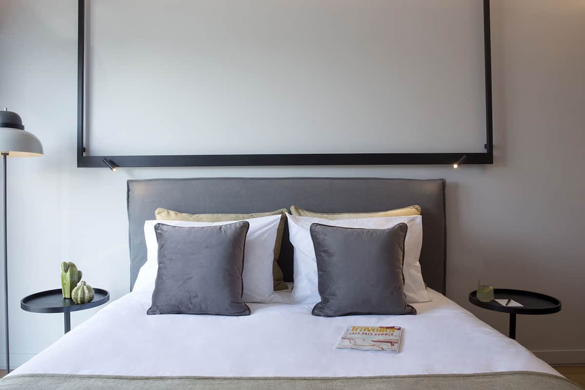 Una habitación de hotel moderna con una cama grande, dos mesitas de noche, una lámpara de pie y una revista sobre la cama.