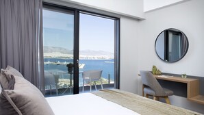 Una habitación de hotel moderna con un gran ventanal corredero que da a un balcón con vistas al mar y a barcos. Hay una cama, un escritorio y una silla en el balcón.