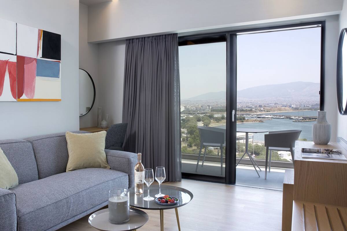 Una sala de estar moderna con un sofá gris, una mesita redonda con una bebida y fruta, y vistas a una ciudad y al agua a través de amplias ventanas.