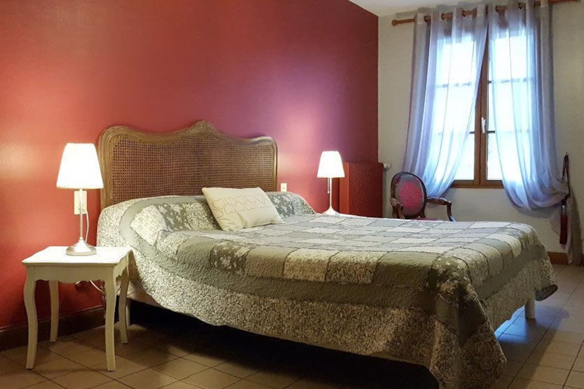 A bedroom with a bed, two bedside lamps, a chair, a window with curtains, and a mirror on the wall.