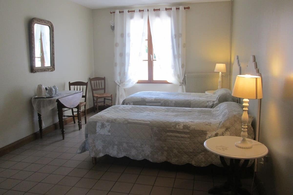 A bedroom with two beds, a small table, a chair, a lamp, and a window with curtains.