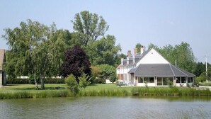 A large house with a covered porch, a pond in the foreground, and trees in the background. - Le Champ du Pre (Gievres)