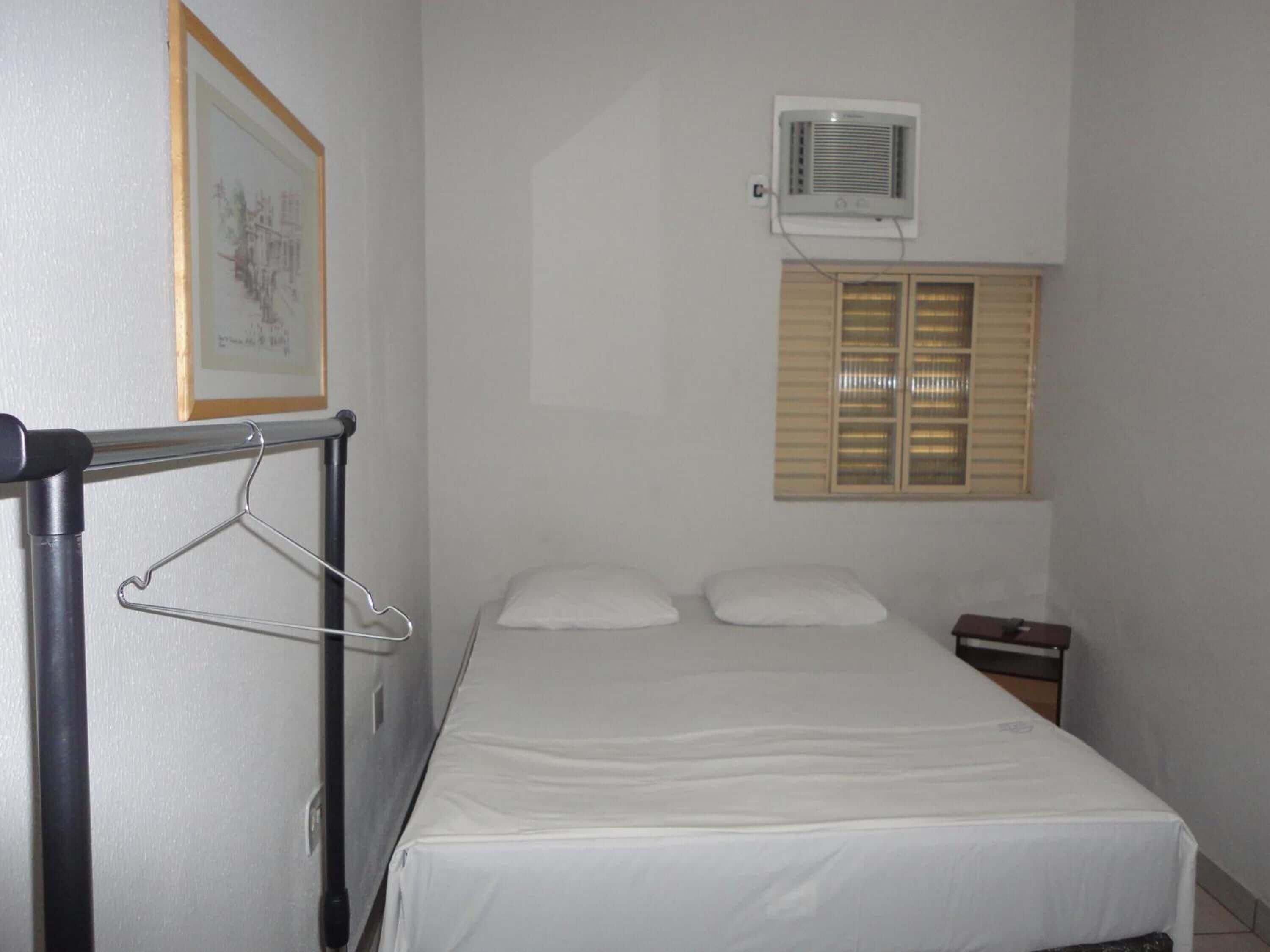 A single bed with a white mattress and two pillows, a small bedside table, a framed picture on the wall, and a window with blinds.