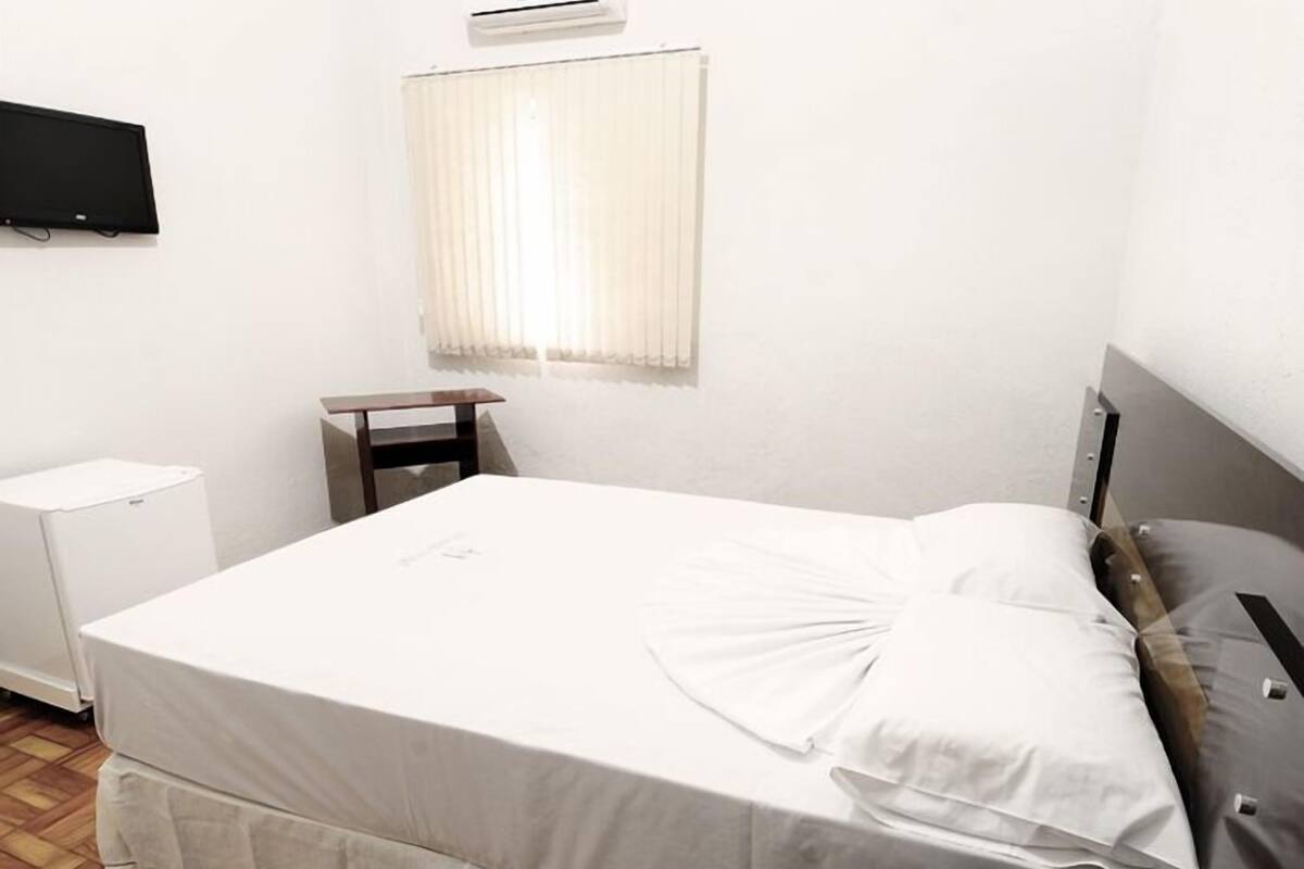 Una cama bien hecha con edredón y almohadas blancos, una mesita de noche, un televisor montado en la pared y una ventana con persianas.