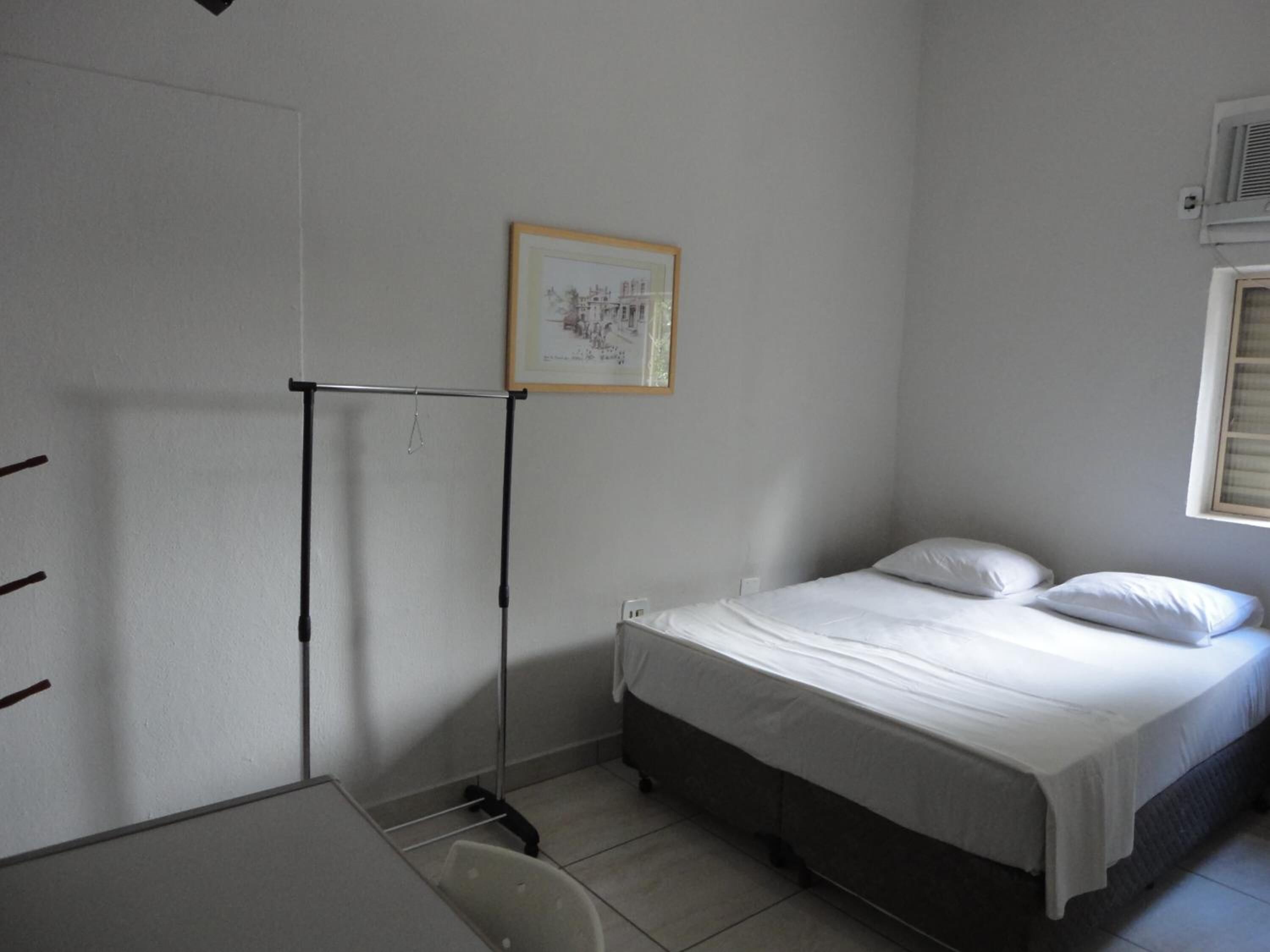 A small, clean room with a single bed, a desk, and a chair. There is a framed picture on the wall and a window with blinds.