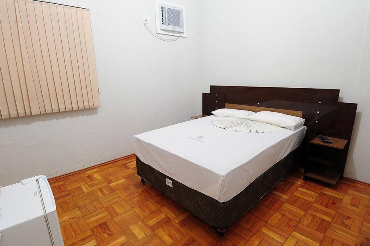 Una cama individual con cabecera de madera, una mesita de noche, una pared blanca, un radiador y un suelo de parqué de madera.