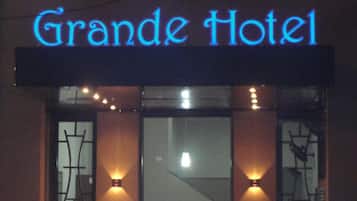 Fachada del Hotel Grande iluminada de noche, con señalización brillante, amplias ventanas con vidrieras decorativas y dos plantas en macetas en la planta baja.