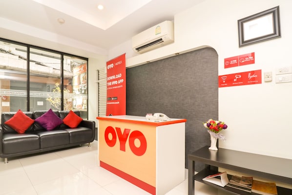A hotel reception area with a reception desk labeled "OYO," a black leather sofa with red and purple cushions, a vase with flowers, and a wall-mounted air conditioner.