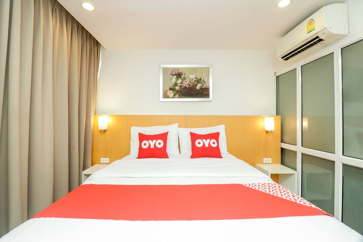 Une chambre d’hôtel avec un lit, deux oreillers sur lesquels est inscrit le mot « OYO », un climatiseur fixé au mur, un tableau au-dessus du lit et une fenêtre avec des stores.