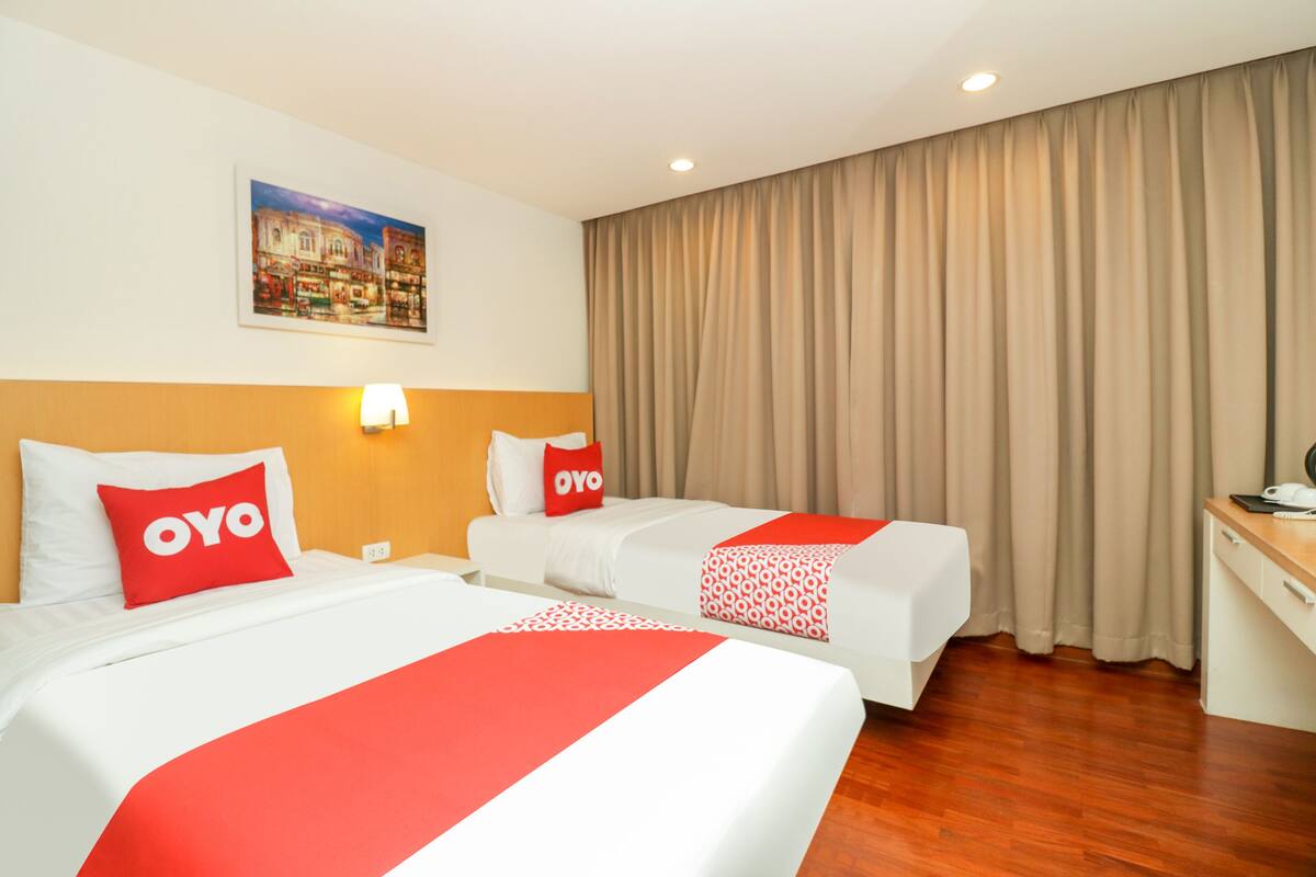 Une chambre d’hôtel avec deux lits, chacun recouvert d’une couvre-lit rouge et blanche, une tête de lit ornée d’un tableau représentant un paysage urbain, un bureau avec une lampe et une grande fenêtre avec des rideaux.