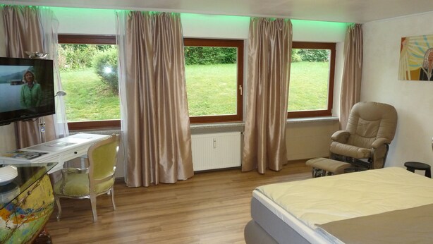 A hotel room with a bed, a desk with a chair, a TV, and large windows with curtains. - Ferienwohnung Jeremy (Bad Toelz)