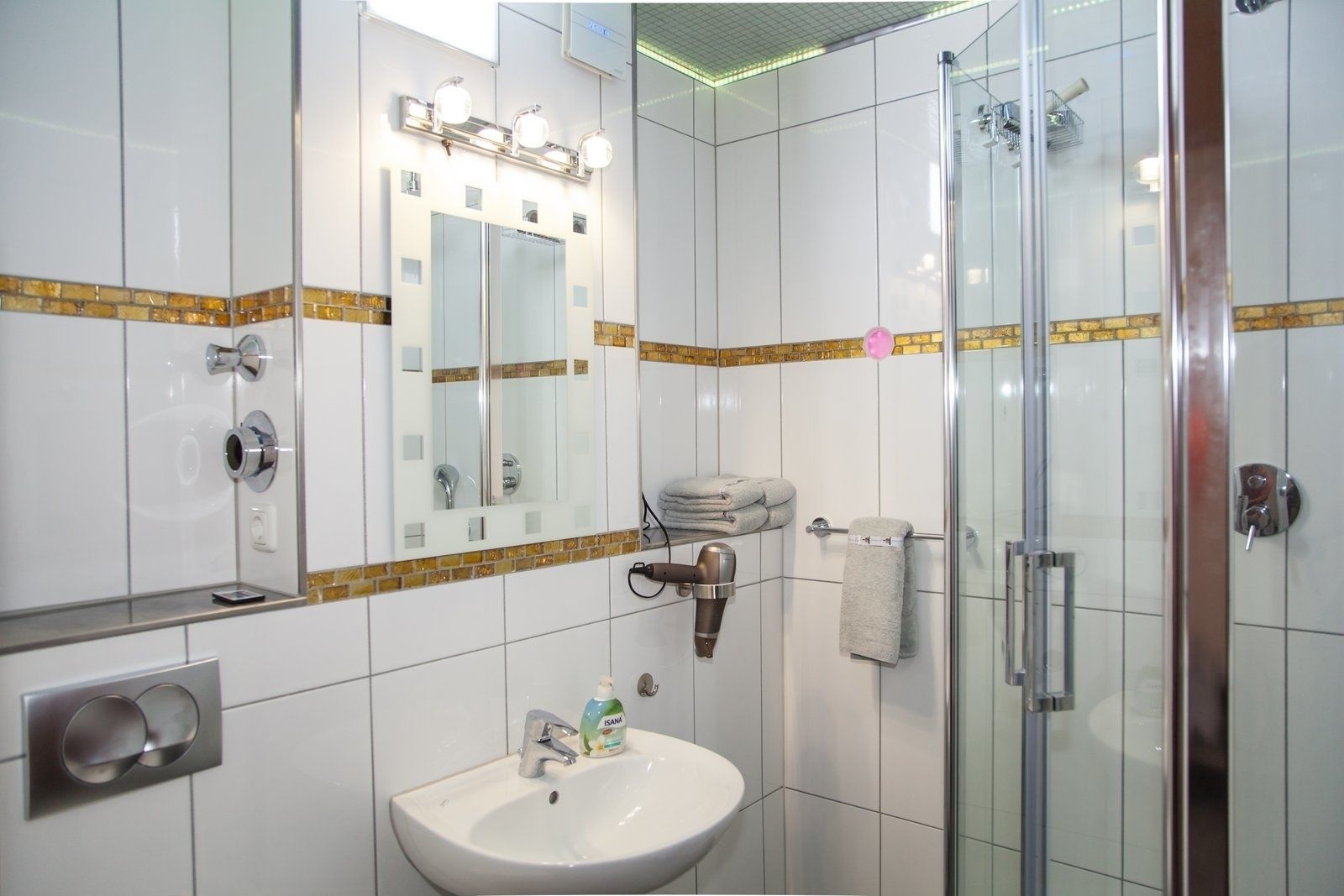 A modern bathroom with a glass shower enclosure, a sink with a soap dispenser, a towel rack, and a mirror with lights above it.