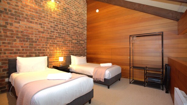 Habitación de hotel con dos camas, una pared de ladrillo, paneles de madera y un perchero metálico.